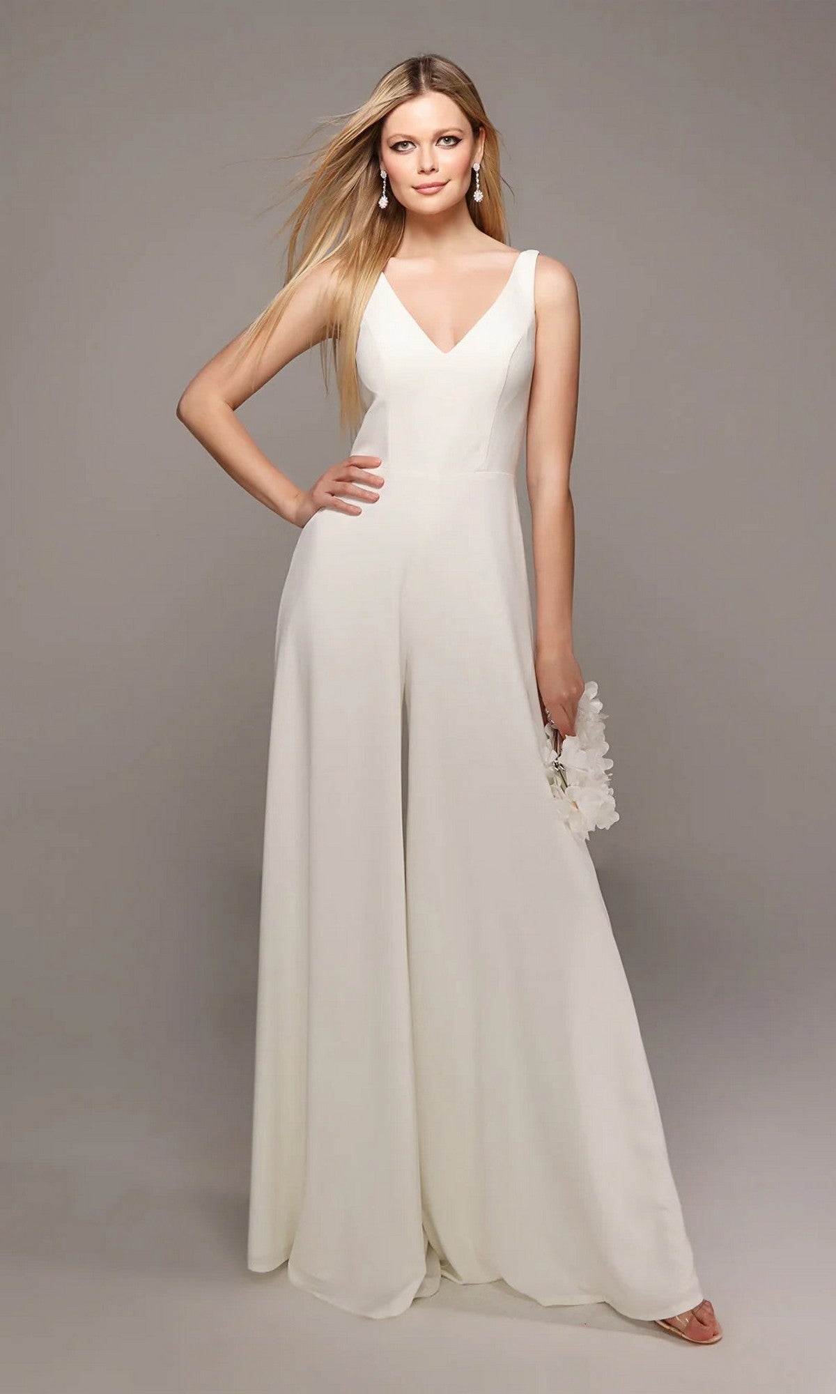 A woman stands against a plain grey background wearing the Long White Jumpsuit: Alyce 70021 with wide-leg pants, holding a small white clutch.